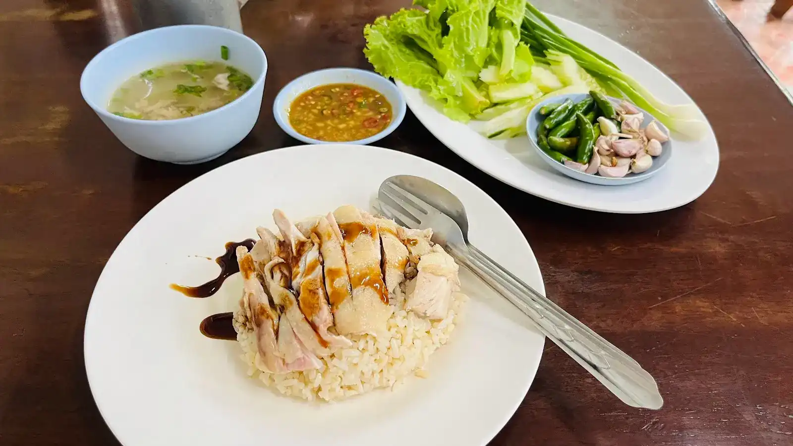 Hainanese chicken rice with dark sauce, a bowl of clear soup, dipping sauce and a plate of greens, chillies and garlic
