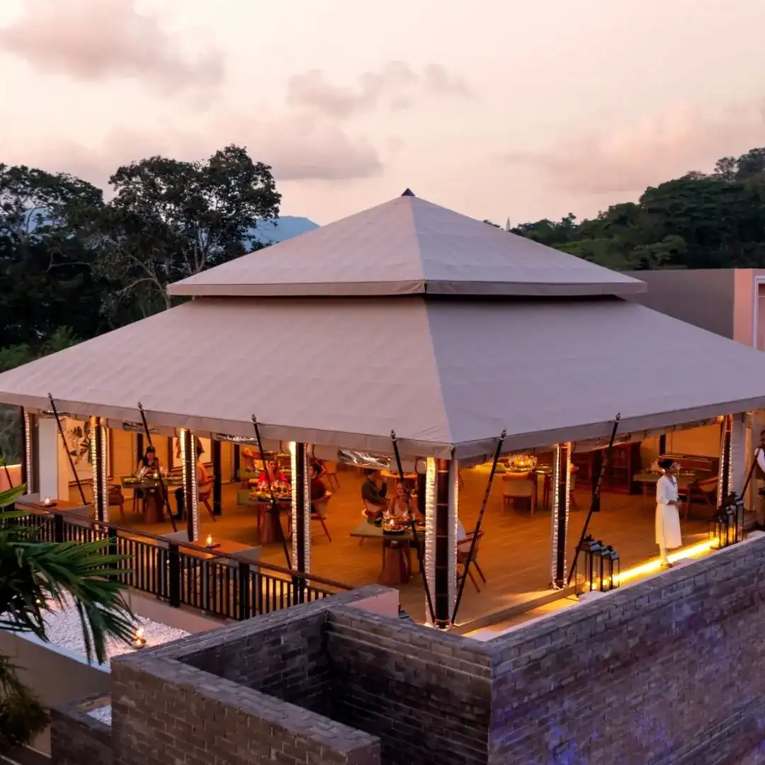 The two-tiered tent roof of Tambu at dusk, tables lit underneath, forest hills behind