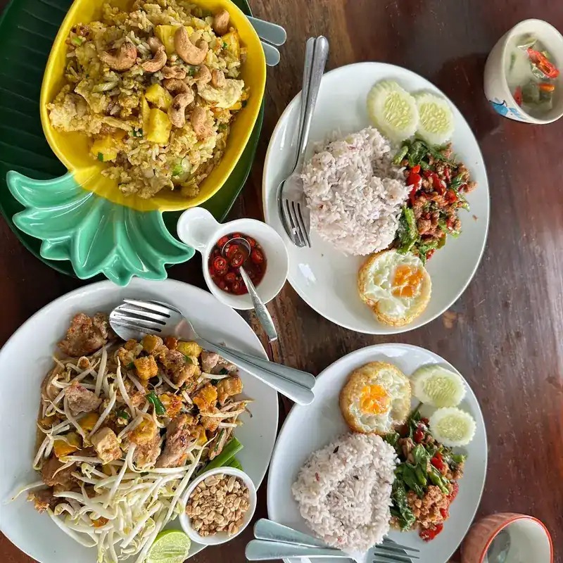 A table seen from above: fried rice in a pineapple-shaped bowl, pad thai with peanuts and lime, and two stir-fries on rice with fried eggs