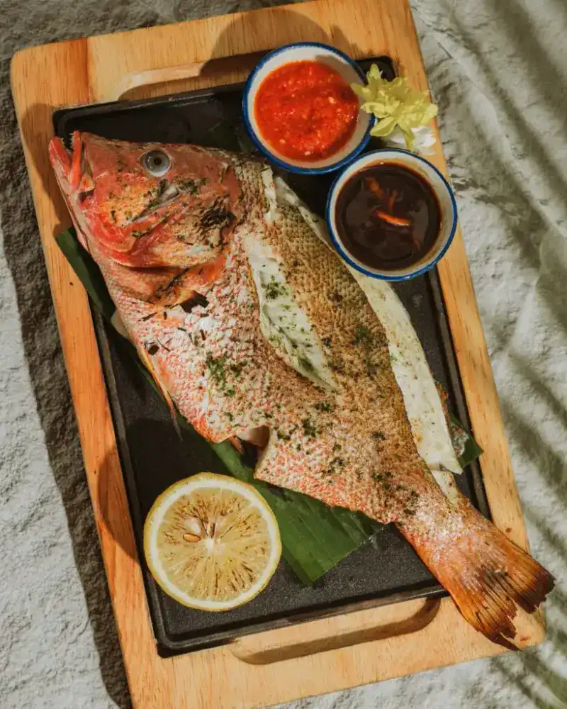 A whole charcoal-grilled red snapper on a wooden board with lemon and two dipping sauces