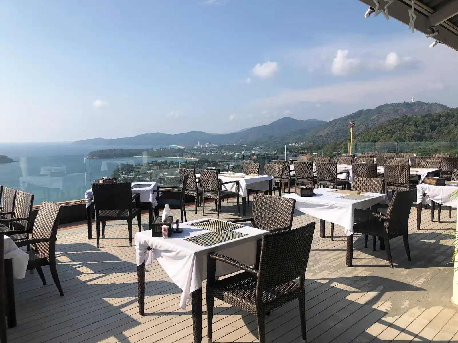 The open deck at Heaven: rattan chairs, white tablecloths, the sea and the bays below / ระเบียงเปิดโล่งของร้าน Heaven เก้าอี้หวาย โต๊ะปูผ้าขาว ทะเลและอ่าวเบื้องล่าง