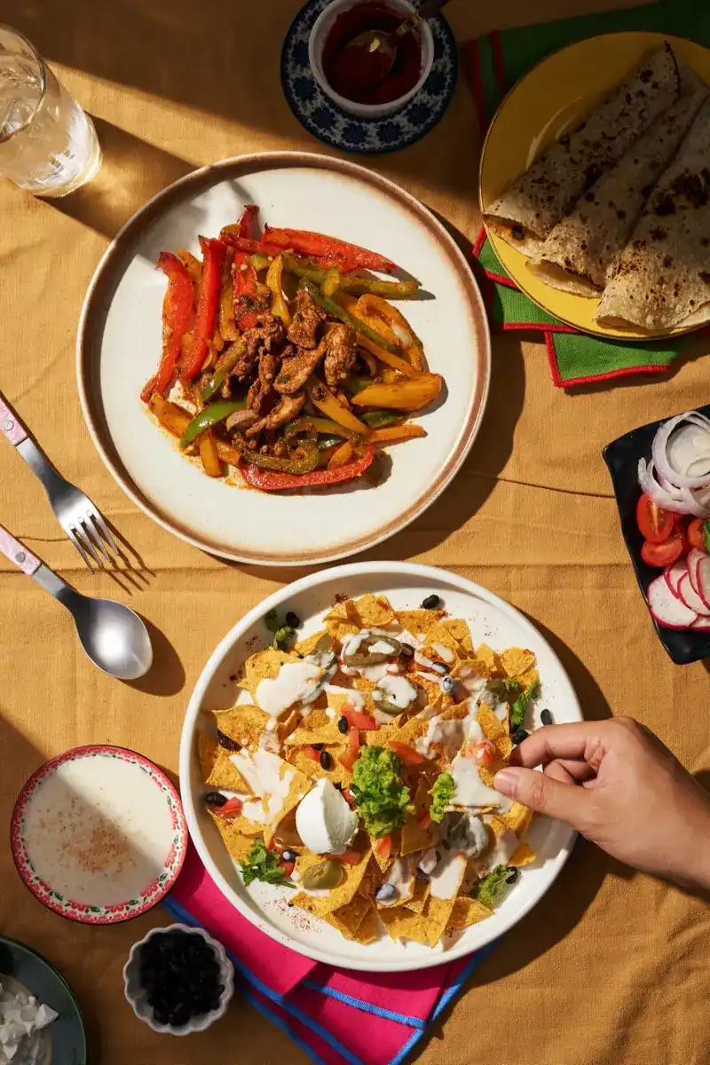 Fajitas, loaded nachos and warm tortillas on a mustard tablecloth, seen from above