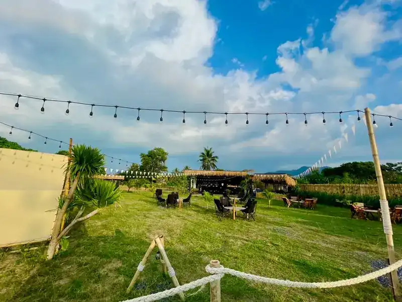 The yard by day: lawn, camping tables, thatched huts and strings of lights under a big sky