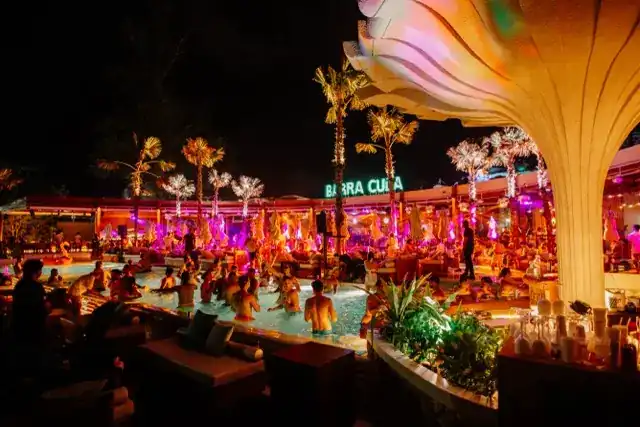 The pool after dark, with palms lit in pink and gold and the club full of guests