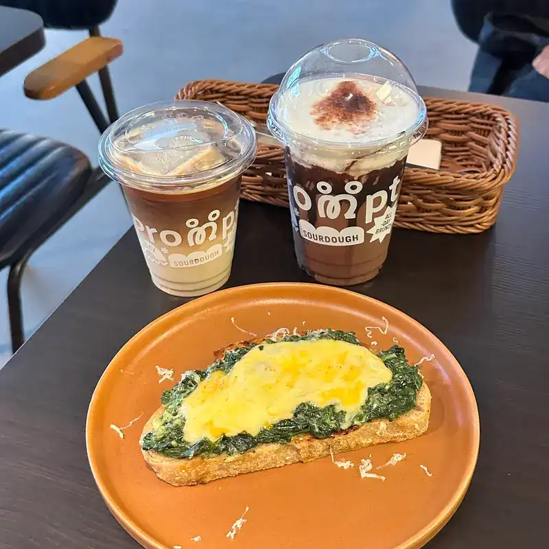 Open sourdough with spinach and melted cheese on an orange plate, with two iced drinks in PROMPT cups