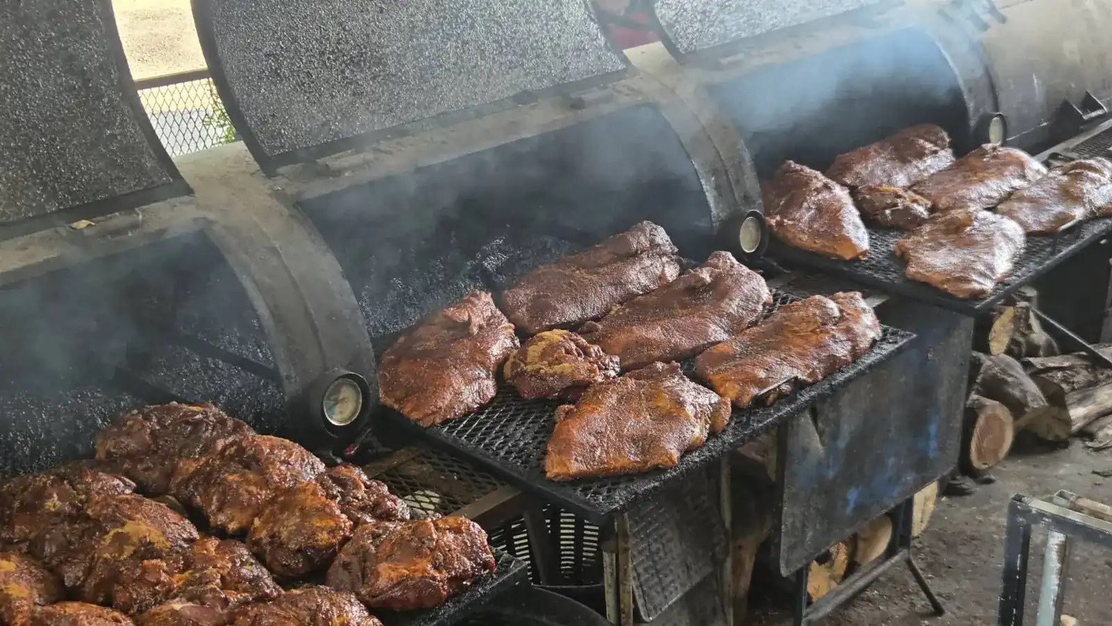 Pork ribs and pork shoulders smoking on the open barrel smokers, with firewood stacked beside them