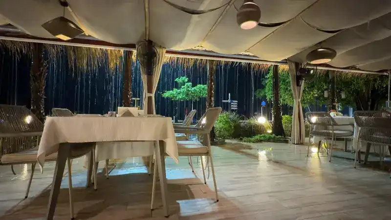 The terrace on a rainy night: lamps lit under the canvas, a table laid in white, rain falling past the palms