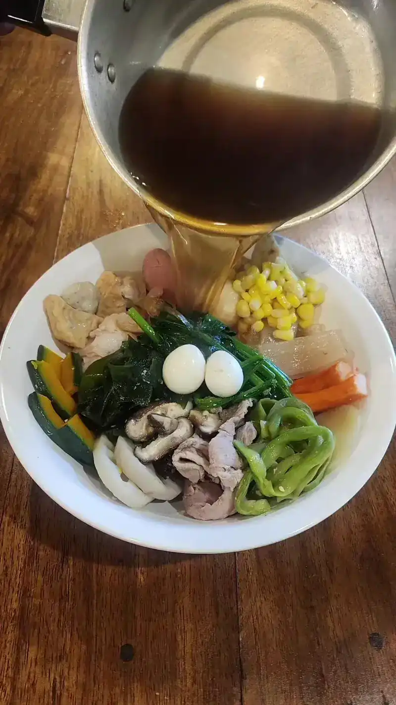 Hot broth being poured over a bowl of greens, quail eggs, sweetcorn, mushrooms and sliced meat.