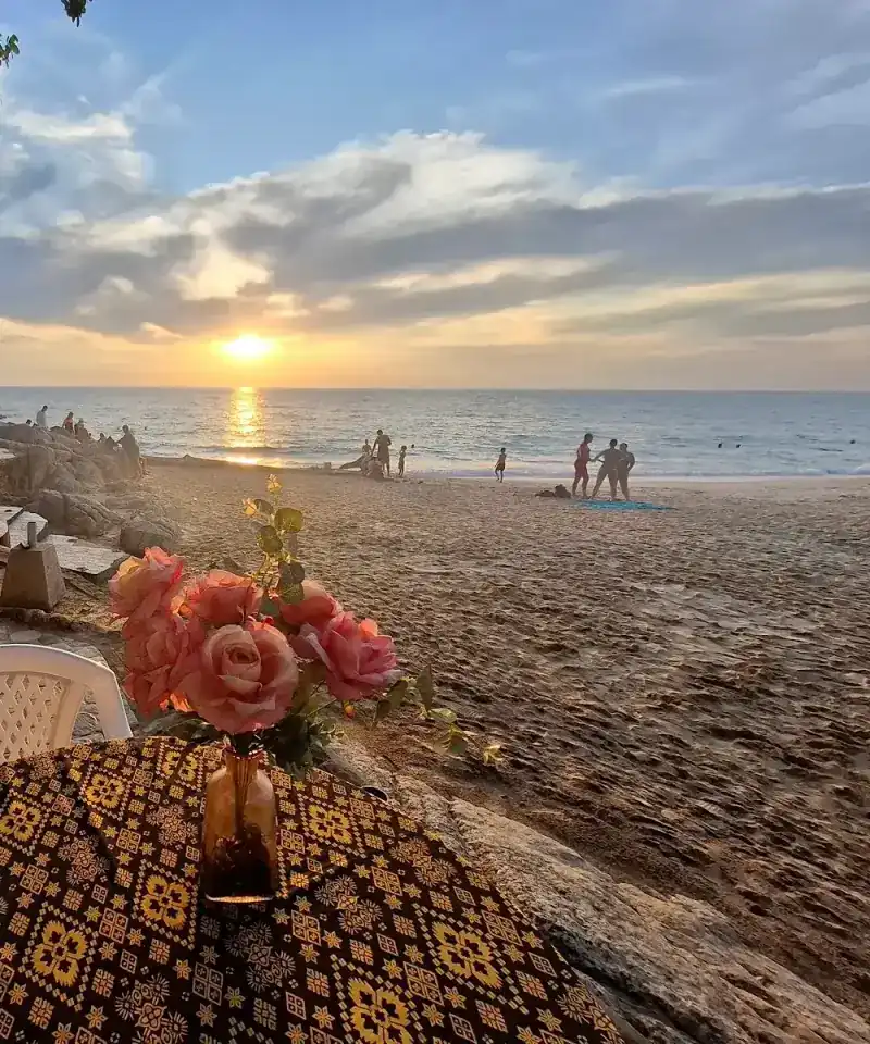 Sunset over the sea seen from a table with pink roses and a black-and-gold cloth, people walking on the sand.