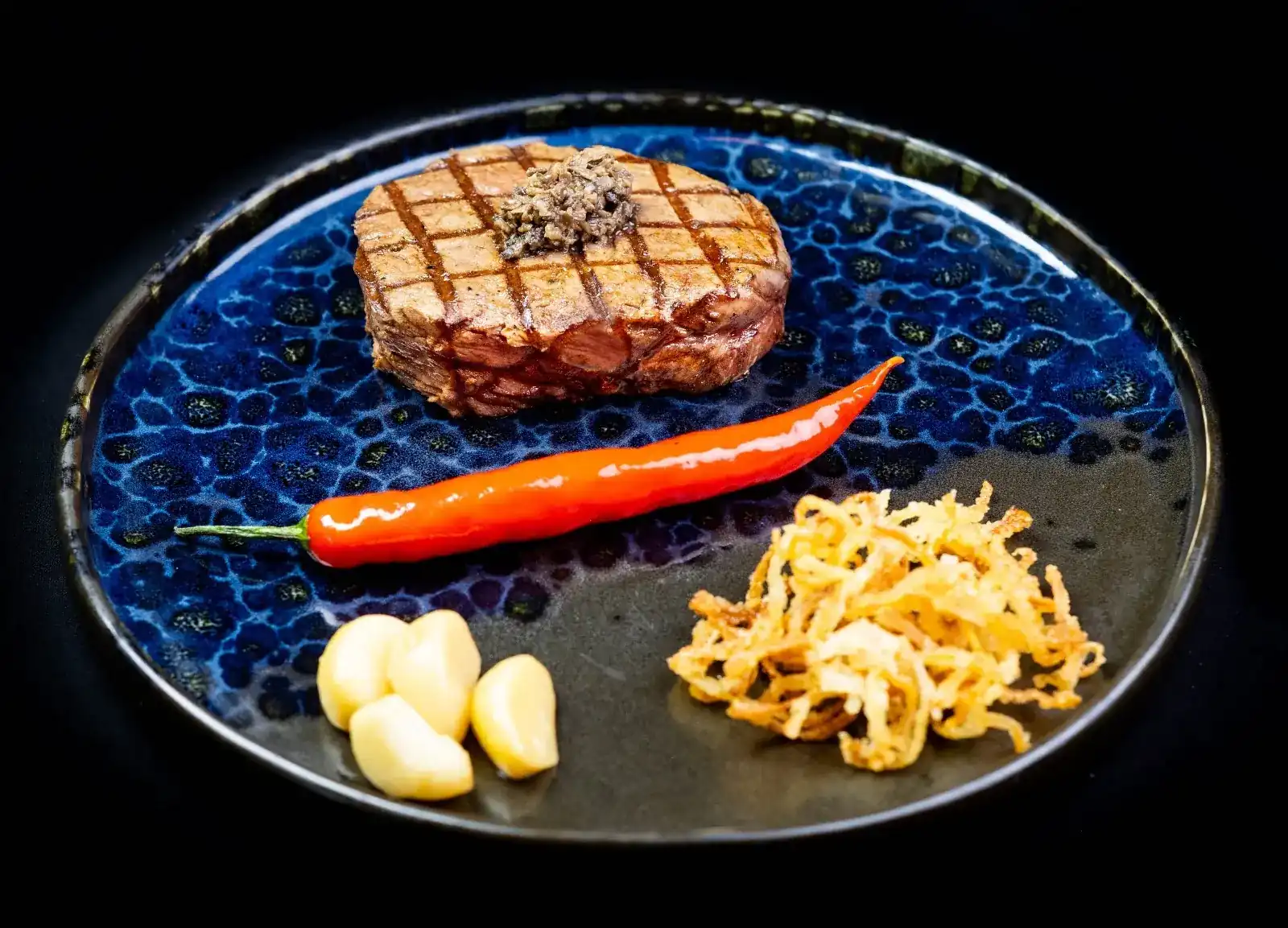 Char-grilled steak with crosshatch grill marks on a blue glazed plate, with a red chili, roasted garlic and crispy onions