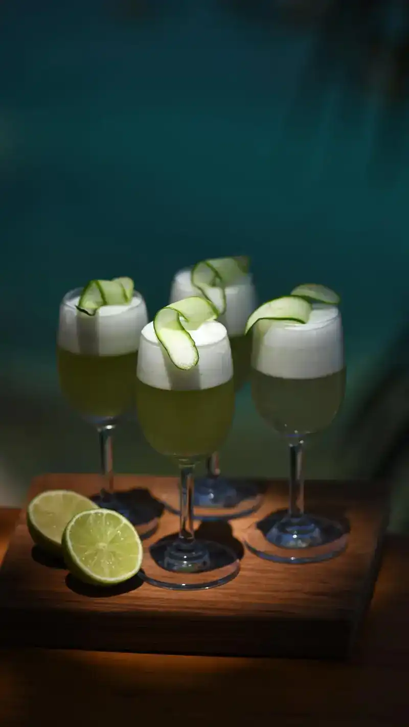 Four foamy green drinks with cucumber ribbons and cut limes