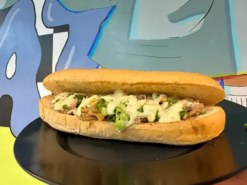 A long filled roll with spring onion and a creamy sauce, on a black plate in front of the painted wall