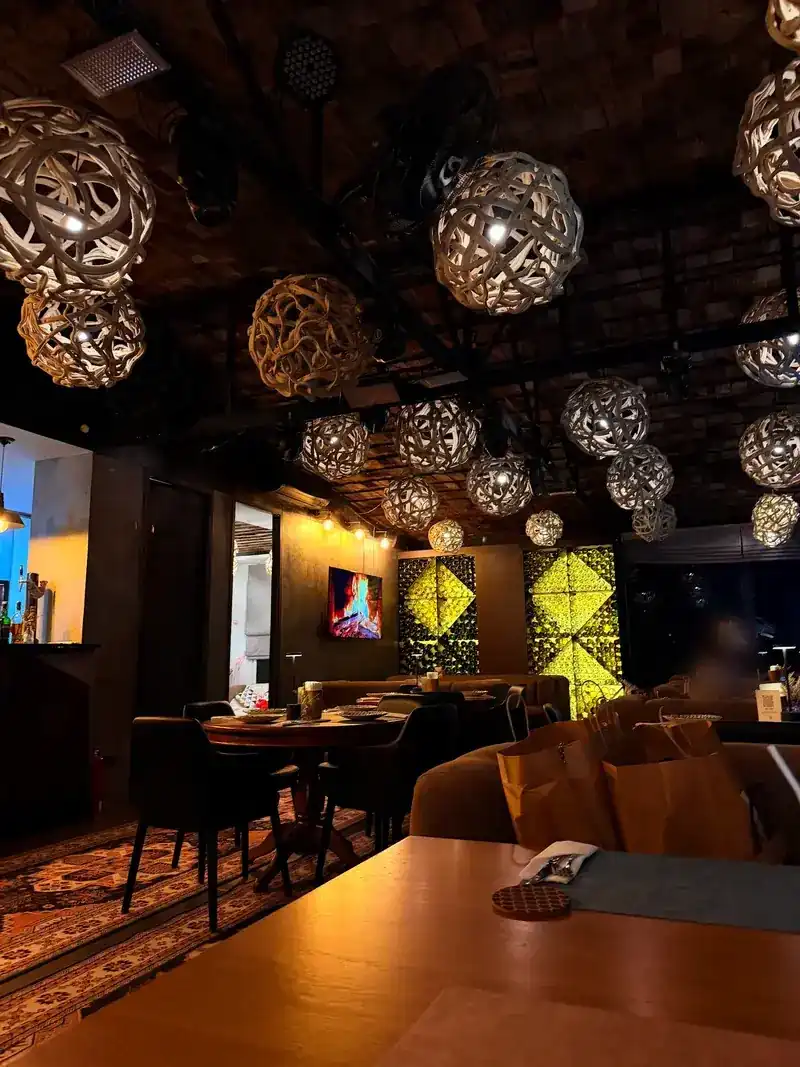 The dining room inside SALTY: woven ball lamps over round tables, dark chairs, a patterned rug and a lit green wall