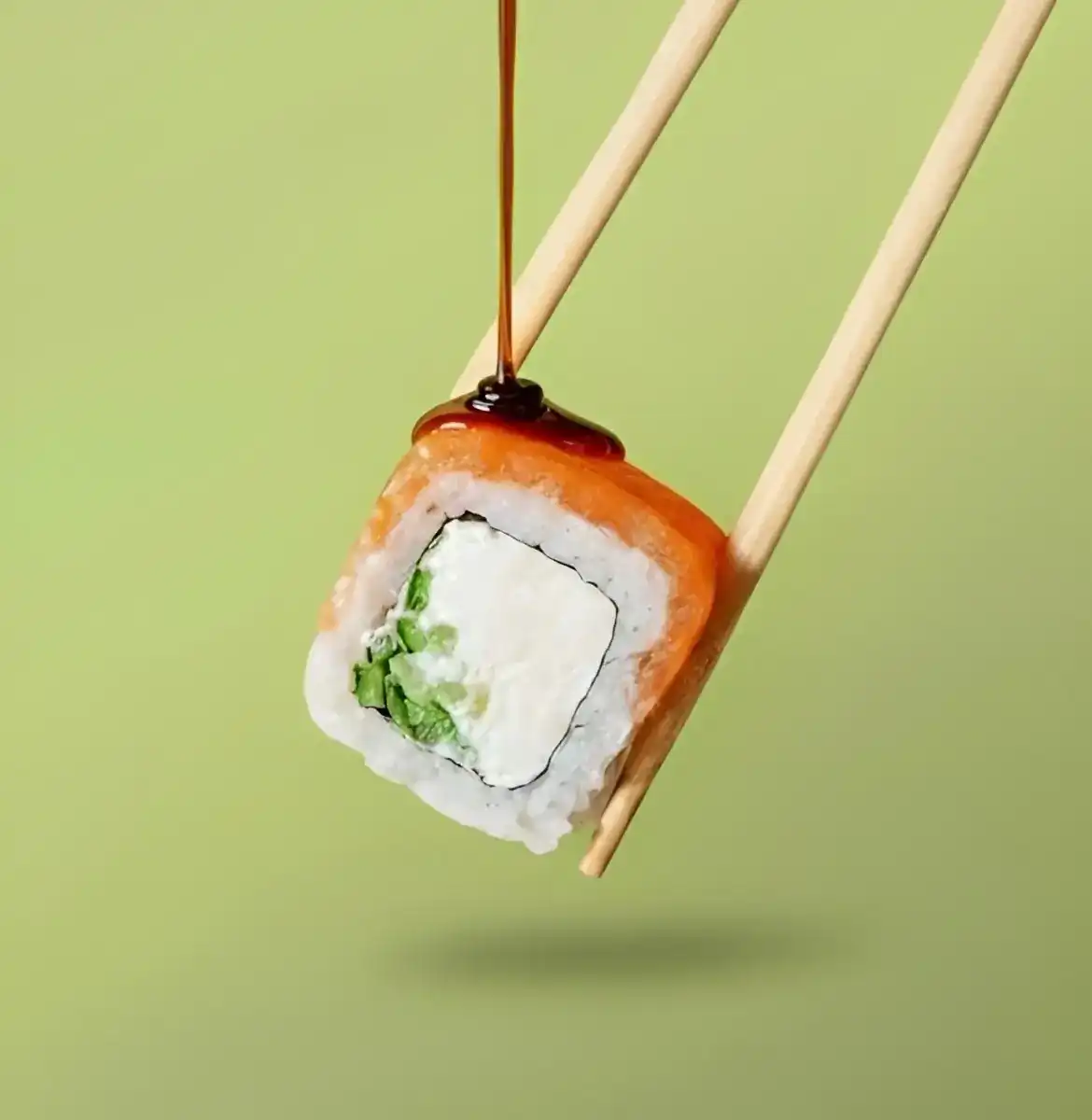 A salmon roll with cream cheese and cucumber held in chopsticks, sauce dripping onto it, on a green background