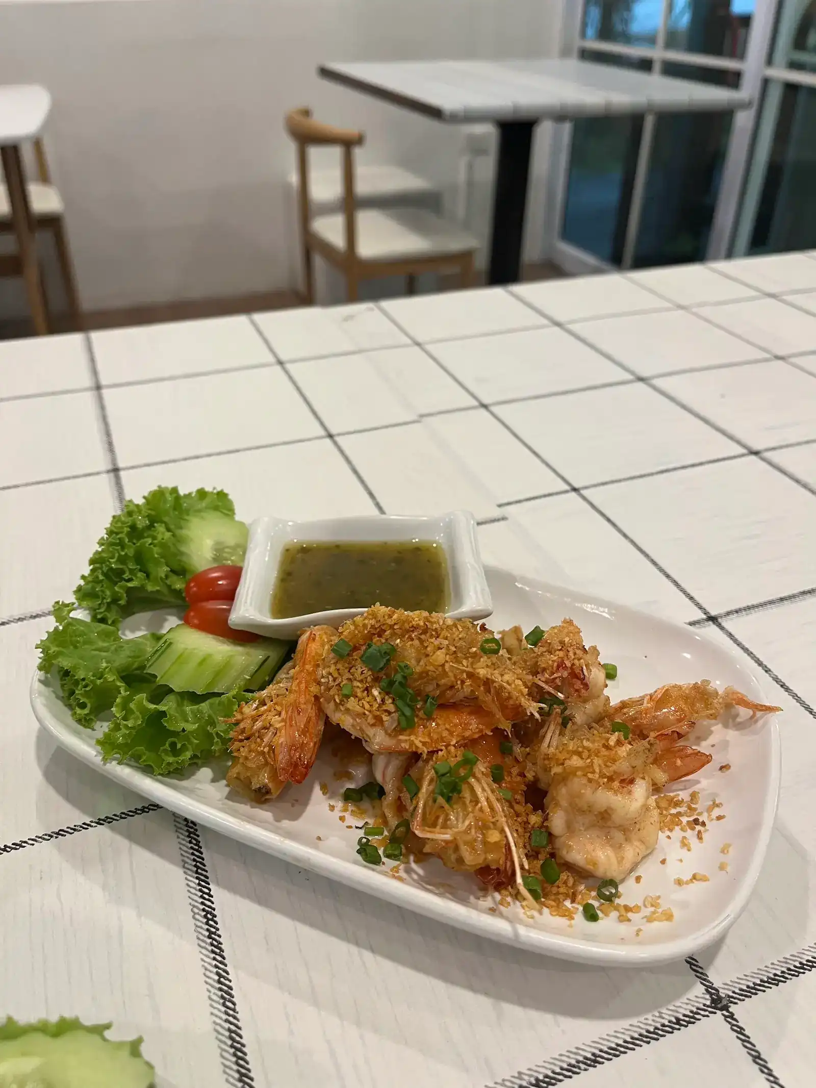 Fried prawns topped with crispy garlic and spring onion, with a green dipping sauce and fresh salad, on a white tiled table