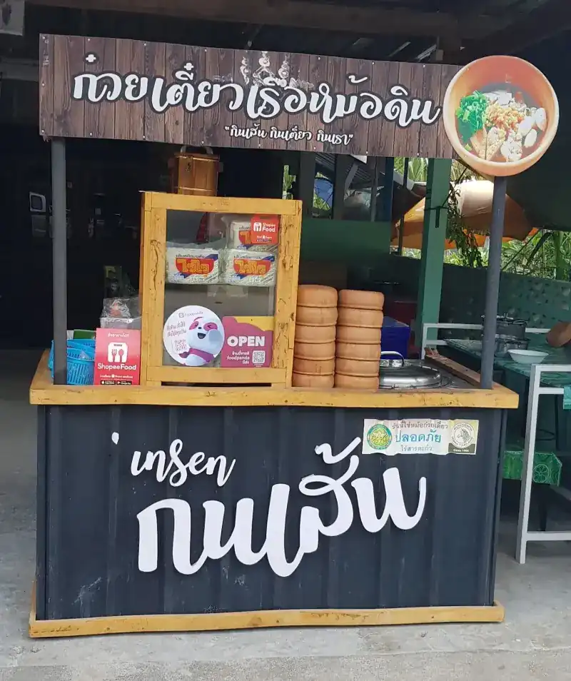 The noodle stall: a wooden sign with the shop's name above a dark cart, clay bowls stacked on the counter