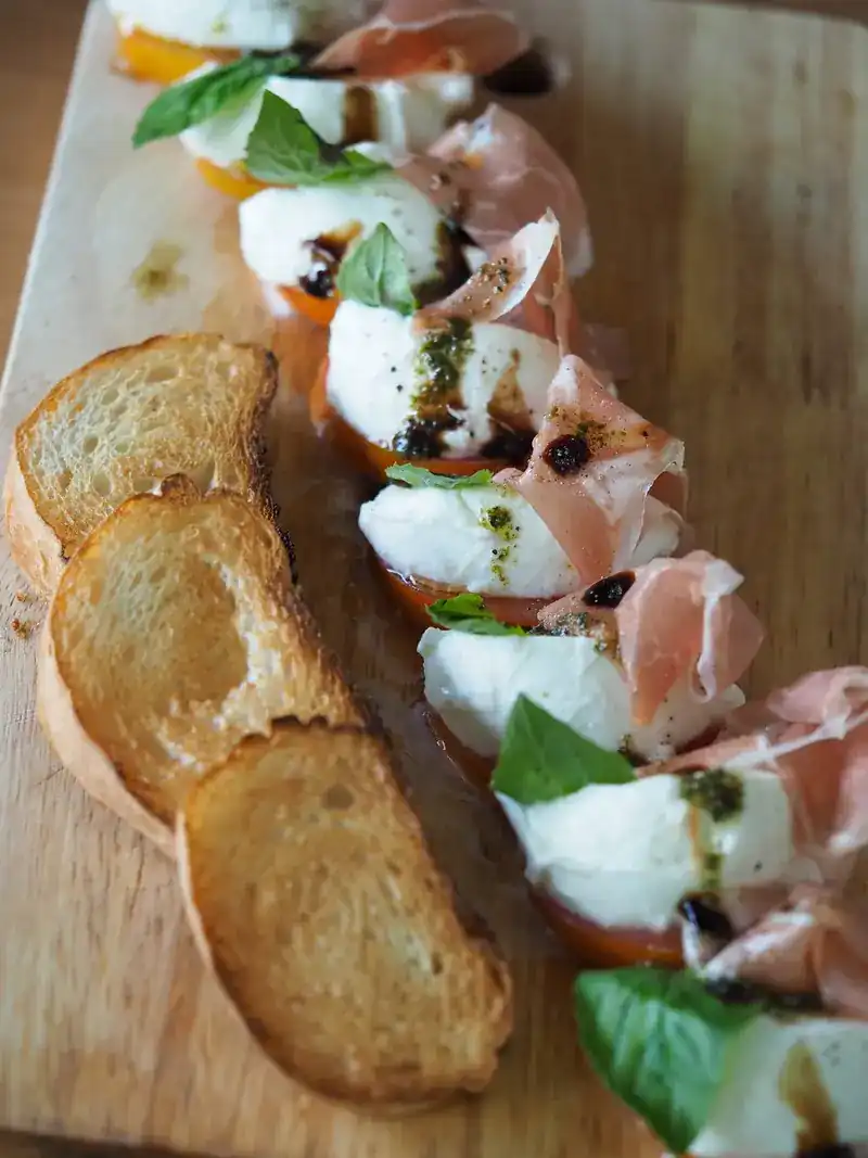 A wooden board with fresh white cheese on tomato, cured ham, basil and toasted bread
