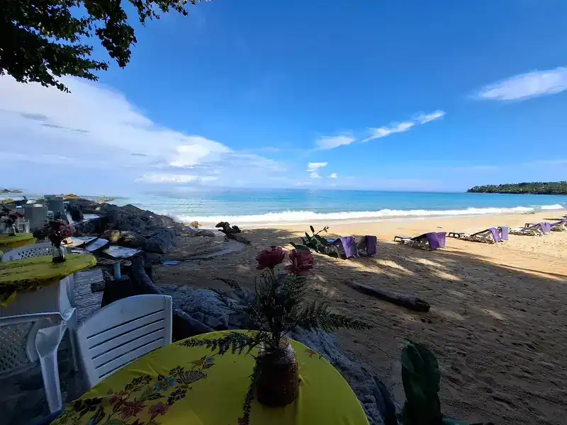 A table with a yellow cloth and flowers at the edge of the beach, rocks and sun loungers beyond, under a blue sky.