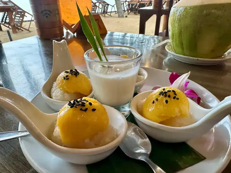 Mango sticky rice served in porcelain spoons, with a young coconut behind